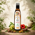 A bottle of Vritikalic 100% Natural Hair Oil stands on a rustic wooden surface surrounded by botanical elements like green grapes, a red hibiscus flower, nuts, and leaves, with soft leaf shadows on a light beige background.